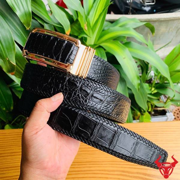 Hình ảnh thắt lưng da cá sấu đan viền TMA9B4, màu đen với khóa vàng, cầm trên tay trước nền lá xanh tươi.