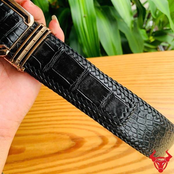 Hình ảnh thắt lưng da cá sấu đan viền TMA9B4 màu đen, khóa vàng, cầm trên tay trước nền lá xanh tự nhiên.