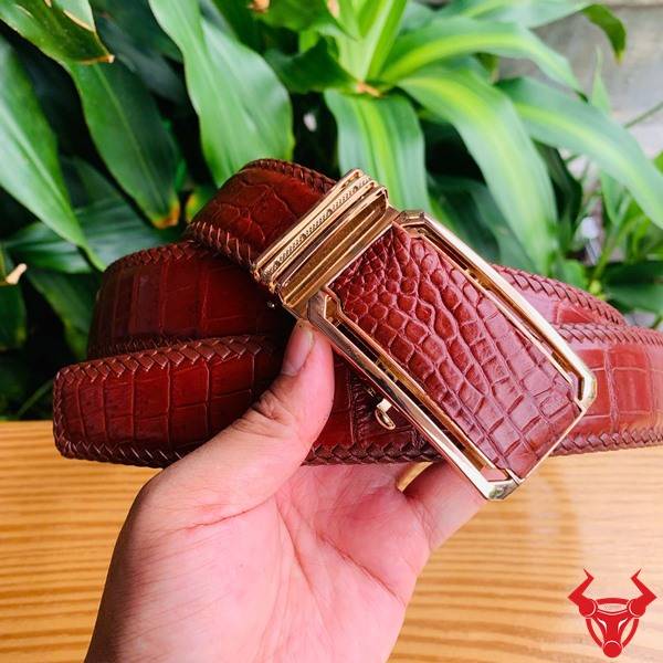 Hình ảnh thắt lưng đan viền da cá sấu nâu đỏ TMA9B4, khóa vàng, cầm trên tay giữa cảnh lá xanh và bàn gỗ.