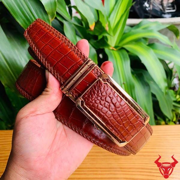 Hình ảnh thắt lưng đan viền da cá sấu nâu đỏ TMA9B4, khóa vàng, cầm trên tay trước nền lá xanh tươi.
