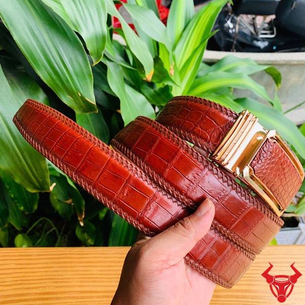 Hình ảnh thắt lưng đan viền da cá sấu nâu đỏ TMA9B4, khóa vàng, cầm trên tay giữa lá xanh và bàn gỗ.