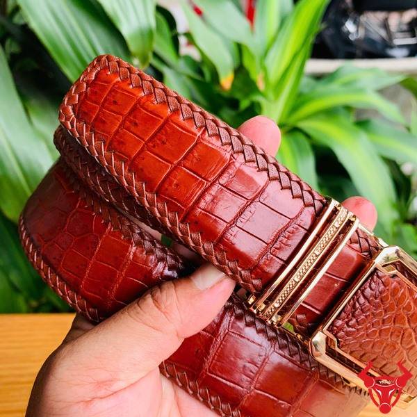 Hình ảnh thắt lưng đan viền da cá sấu nâu đỏ TMA9B4, khóa vàng, cầm trên tay trước nền lá xanh tự nhiên.