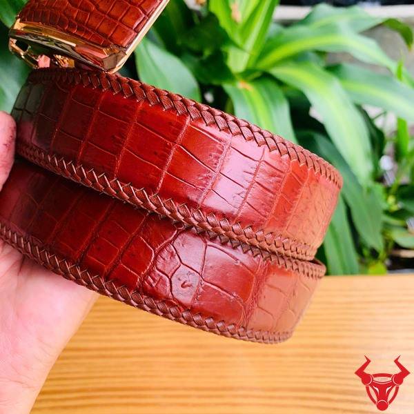 Hình ảnh thắt lưng đan viền da cá sấu nâu đỏ TMA9B4, khóa vàng, cầm trên tay giữa lá xanh và bàn gỗ.