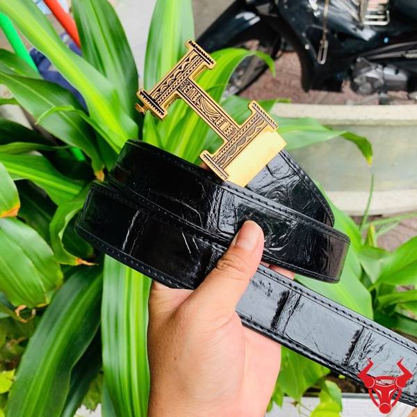 Hình ảnh thắt lưng da cá sấu TSK103, khóa thép mạ vàng, cầm trên tay giữa cảnh lá xanh tự nhiên.