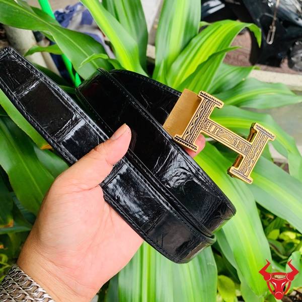 Hình ảnh thắt lưng da cá sấu TSK103, màu đen với khóa thép mạ vàng, cầm trên tay trước nền lá xanh.