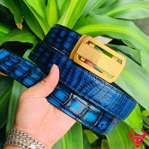 Hình ảnh thắt lưng da cá sấu xanh patina TSK33, khóa vàng, cầm trên tay giữa cảnh lá xanh và nền tự nhiên.