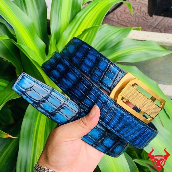 Hình ảnh thắt lưng da cá sấu xanh patina TSK33, khóa thép mạ vàng, cầm trên tay trước nền lá xanh tự nhiên.