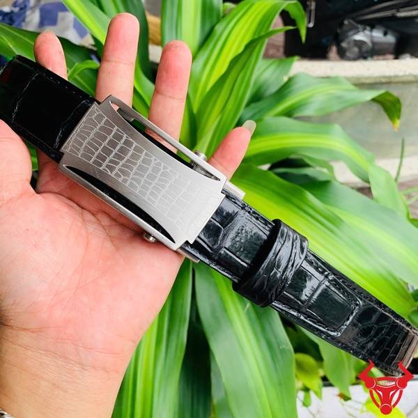 Hình ảnh thắt lưng da cá sấu TSK32, khóa lăn inox họa tiết cá sấu, cầm tay trên nền lá xanh.