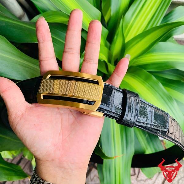 Hình ảnh thắt lưng da cá sấu TSK33, khóa tỳ thép vàng, cầm trên tay với lá xanh.