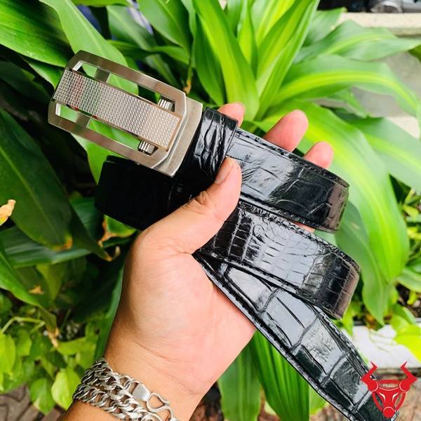 Hình ảnh dây lưng da cá sấu TSK33, khóa tỳ thép mạ bạc, cầm tay với nền lá xanh.