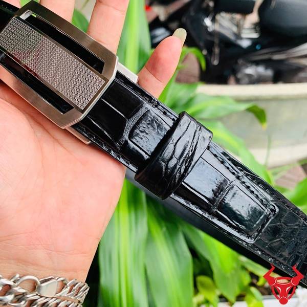 Hình ảnh dây lưng da cá sấu TSK33, khóa tỳ thép mạ bạc, cầm tay trên nền lá xanh.
