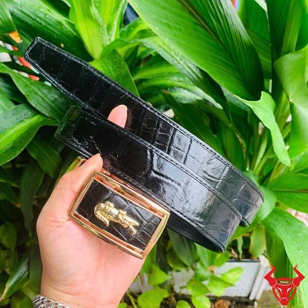Ảnh thắt lưng FBA9B2 da cá sấu đen, khóa vàng con thỏ, được cầm tay với nền thực vật xanh tươi.