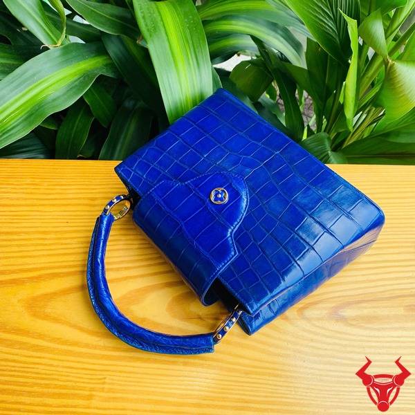 Ảnh túi xách da cá sấu bóng kính màu xanh navy trên bàn, nền lá, cao cấp sang trọng quý phái.