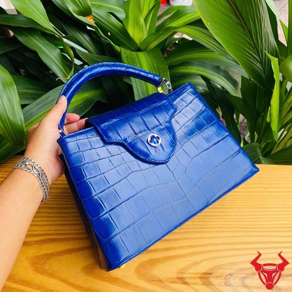 Hình túi xách da cá sấu bóng kính màu xanh navy, nền lá xanh, cao cấp sang trọng quý phái.