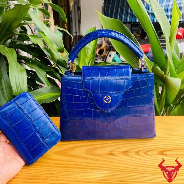 Ảnh túi xách da cá sấu bóng kính màu xanh navy cầm tay, nền lá, cao cấp sang trọng quý phái.