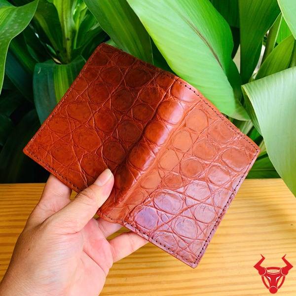 Ảnh ví form đứng VJA8 da cá sấu màu nâu đỏ sáng cầm tay, nền lá, thể hiện cao cấp sang trọng.