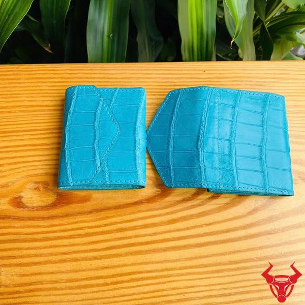 Ba chiếc ví nữ nhỏ gọn da cá sấu xanh ngọc trong hình ảnh, được cầm bởi bàn tay, với nền lá cây xanh, thể hiện phong cách thời trang.