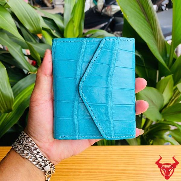 Hình ảnh cầm ba ví nữ da cá sấu mini màu xanh ngọc, họa tiết vân da, nền thực vật xanh, nhấn mạnh phong cách thời trang.