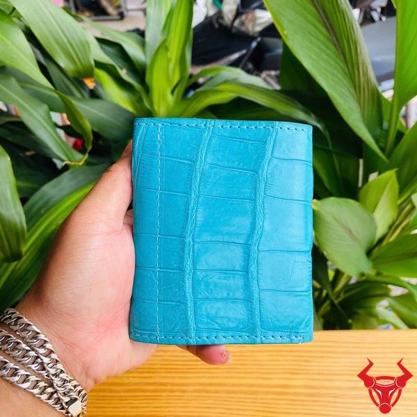 Hình ảnh ba ví nữ nhỏ da cá sấu màu xanh ngọc, họa tiết vân, cầm tay trên nền xanh, thể hiện phong cách thời trang.