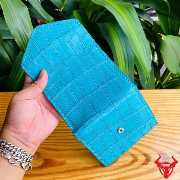 Ba chiếc ví da cá sấu mini xanh ngọc trong hình, được cầm bởi bàn tay, nền lá cây, nhấn mạnh phong cách thời trang.