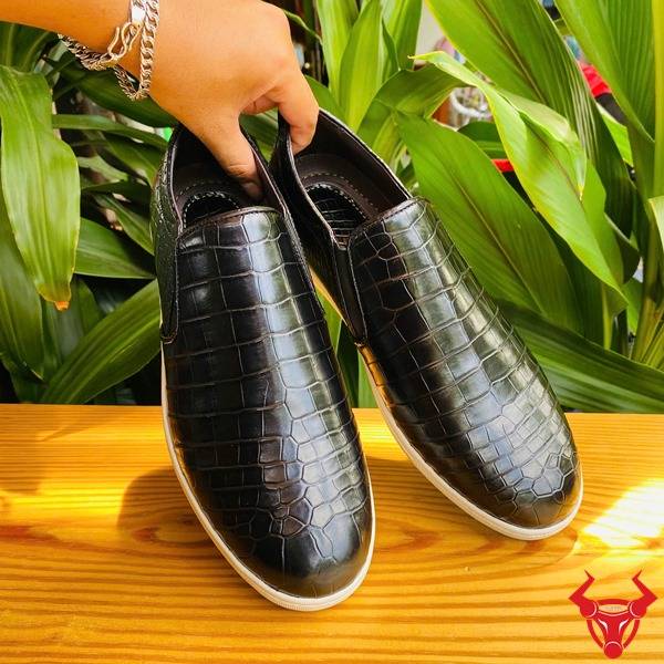 Ảnh đôi giày thể thao da cá sấu đen GCS25, đặt trên bàn với nền lá xanh và logo thương hiệu.