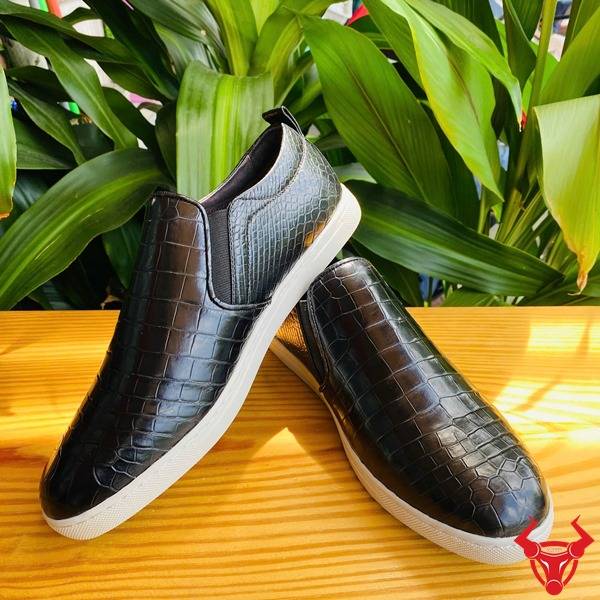 Hình đôi giày thể thao da cá sấu đen GCS25, đặt trên bàn với nền lá xanh và thẻ logo.