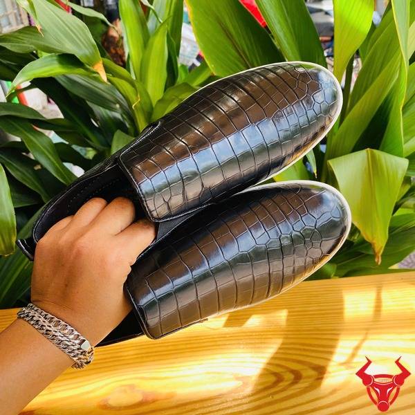 Ảnh chi tiết giày da cá sấu đen GCS25, đặt trên bàn với nền lá xanh và logo.