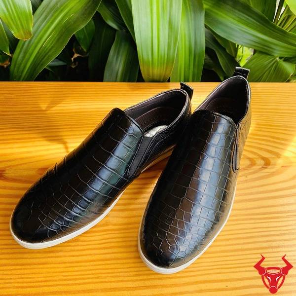 Bức hình giày thể thao da cá sấu đen GCS25, đặt trên bàn với nền lá xanh và thẻ logo.