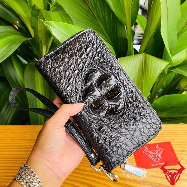 Ảnh ví cầm tay da cá sấu đen lồi, cầm tay với nền lá xanh và logo thương hiệu.
