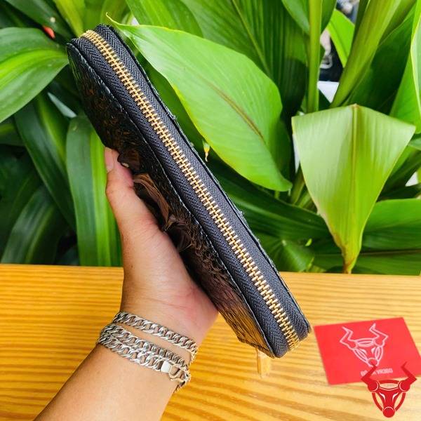 Bức hình ví da cá sấu đen gù lồi, cầm tay với nền lá xanh và thẻ logo.