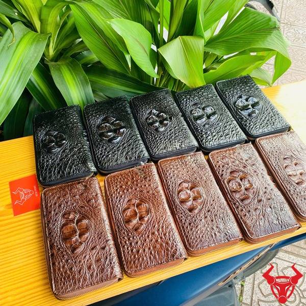 Ảnh ví da cá sấu đen lồi, cầm tay với nền lá xanh và thẻ logo thương hiệu.