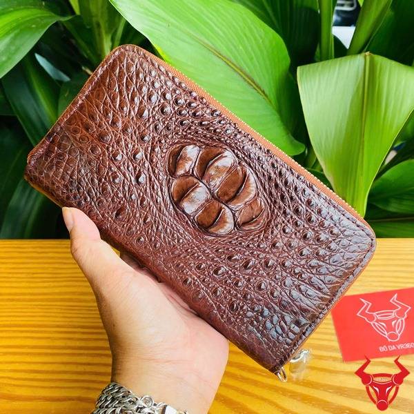 Ảnh sản phẩm ví cầm tay da cá sấu nâu đất lồi, cầm tay với nền lá xanh và logo thương hiệu.