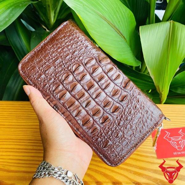 Bức ảnh ví da cá sấu nâu đất gù lồi, cầm tay với nền lá xanh và vòng tay bạc.