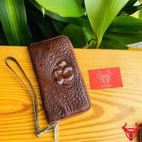 Hình ví da cá sấu nâu đất lồi, cầm tay với nền lá xanh và thẻ logo đỏ.
