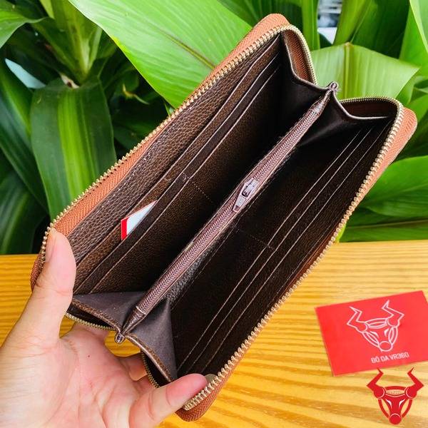 Ảnh chi tiết ví da cá sấu nâu đất lồi, cầm tay trên nền gỗ và lá xanh.