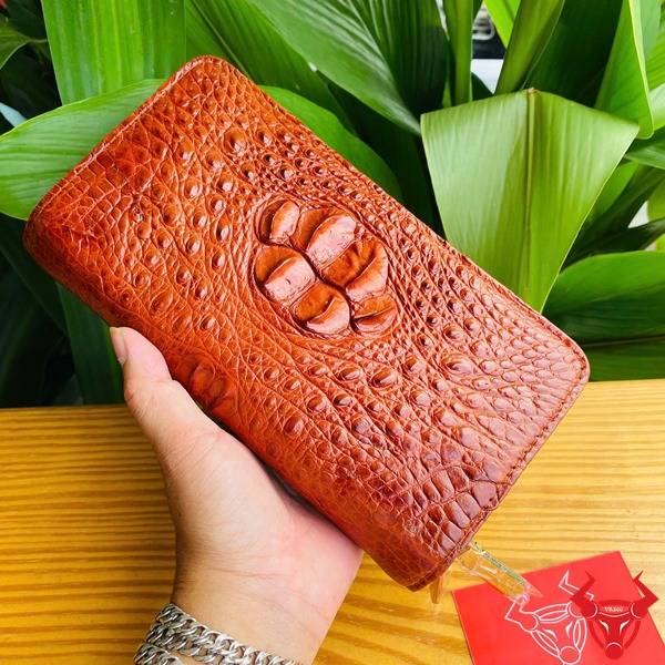 Ảnh sản phẩm ví cầm tay da cá sấu nâu đỏ với 2 khuôn thụt, đặt trên bàn gỗ với nền lá xanh.