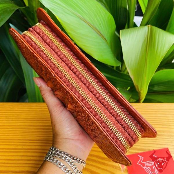 Bức ảnh ví da cá sấu nâu đỏ gù đặt trên bàn, với nền lá xanh và thẻ logo đỏ.