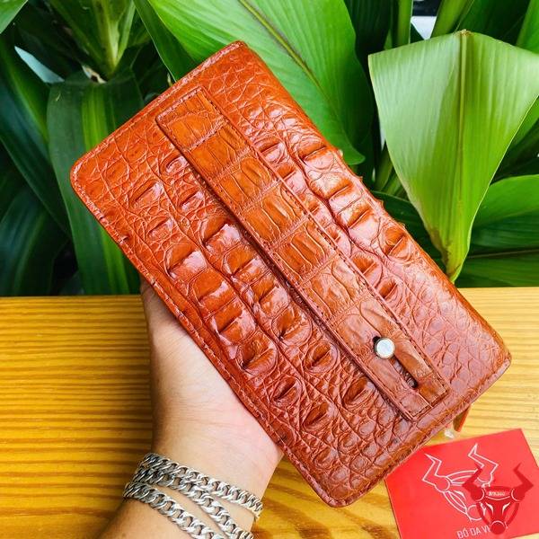 Hình ảnh ví da cá sấu nâu đỏ với họa tiết gù, đặt trên bàn gỗ với lá xanh và thẻ logo.