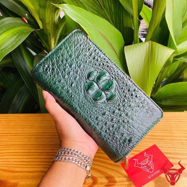 Ảnh sản phẩm ví cầm tay da cá sấu xanh lá với 2 khuôn thụt, cầm tay trên nền thực vật.