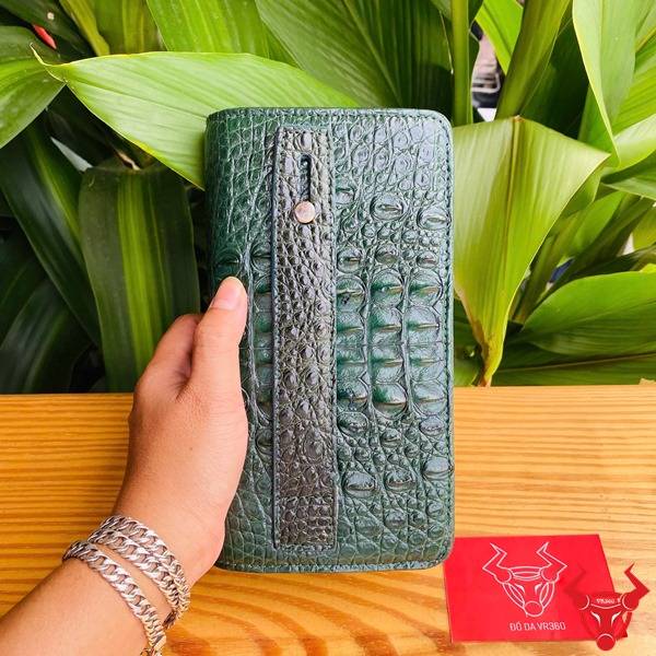 Bức ảnh ví da cá sấu xanh lá gù được cầm, với vòng tay bạc và nền lá xanh.