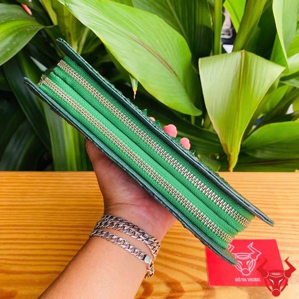Hình cầm ví da cá sấu xanh lá với họa tiết gù, nền lá xanh và thẻ logo đỏ.