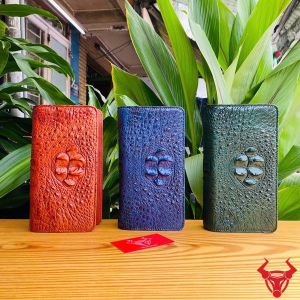 Bức hình ví da cá sấu xanh lá gù được cầm, với nền lá xanh và phụ kiện vòng tay.
