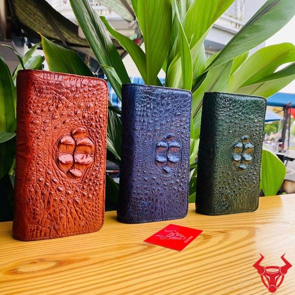 Hình ảnh ví da cá sấu xanh lá gù cầm tay, với nền lá xanh và thẻ logo thương hiệu.
