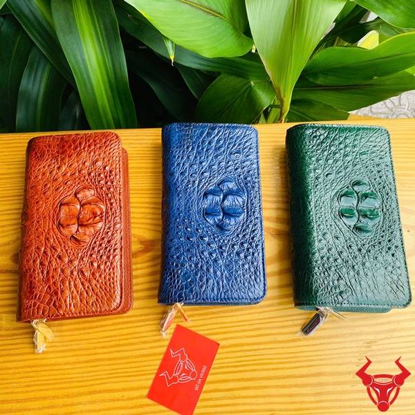 Ảnh ví da cá sấu xanh lá với 2 khuôn thụt, cầm tay bên cạnh thẻ logo đỏ và lá xanh.