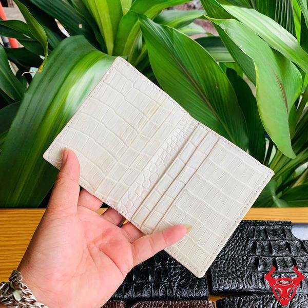 Hình cầm ví da cá sấu trắng vân nổi, nền lá xanh và logo thương hiệu.