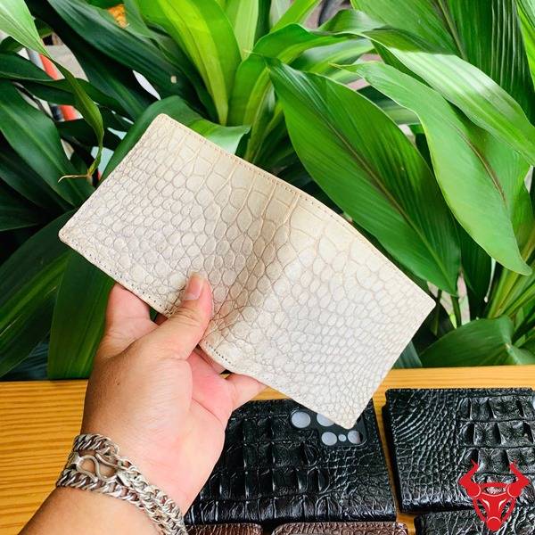Ví Da Bụng Cá Sấu Nam Bạch Tạng Thời Trang Cao Cấp Sang Trọng Bền Bỉ Ảnh sản phẩm ví da cá sấu trắng bạch tạng hai mặt, cầm tay với nền thực vật xanh và logo con bò.