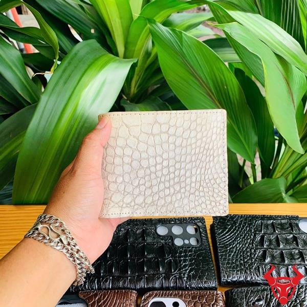 Thời Trang Cao Cấp Sang Trọng Bền Bỉ Ví Nam Da Cá Sấu VCA8 Bạch Tạng Hình ảnh bàn tay cầm ví nam da cá sấu trắng bạch tạng với vân da sang trọng, nền lá xanh và các phụ kiện da khác.