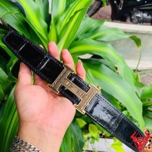 Hình ảnh thắt lưng da cá sấu TSK103, màu đen với khóa thép mạ vàng, được cầm trên tay trước nền lá xanh tươi sáng.