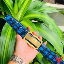 Hình ảnh thắt lưng da cá sấu xanh patina TSK33, khóa thép mạ vàng, được cầm trên tay trước nền lá xanh tươi sáng.