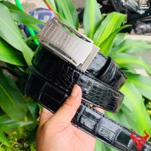 Hình ảnh thắt lưng da cá sấu TSK32 màu đen, khóa lăn inox họa tiết cá sấu, cầm tay trên nền lá xanh.